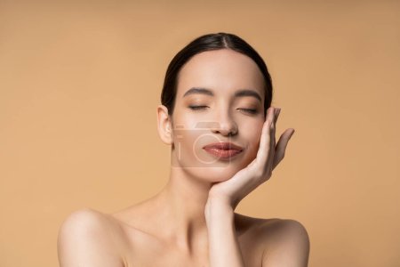 Junge asiatische Frau mit nackten Schultern und geschlossenen Augen berühren Wange isoliert auf beige