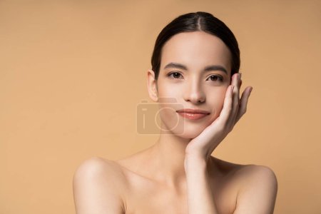 Young asian model with naked shoulders and perfect skin touching cheek isolated on beige