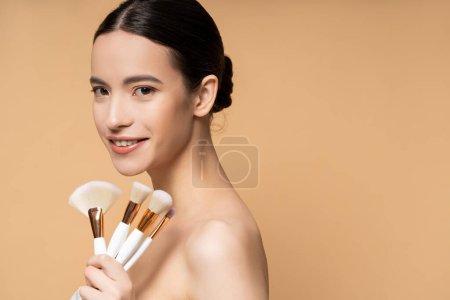 Smiling asian woman with naked shoulders holding makeup brushes and standing isolated on beige