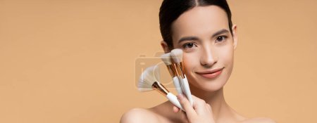 Smiling asian model with naked shoulders holding makeup brushes isolated on beige, banner