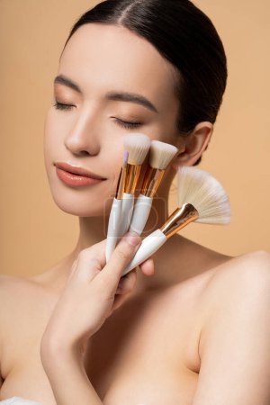 Pretty asian woman with naked shoulders holding makeup brushes and closing eyes isolated on beige