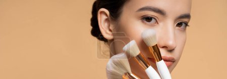 Joven mujer asiática mirando a la cámara y la celebración de cepillos de maquillaje cerca de la cara aislado en beige, pancarta