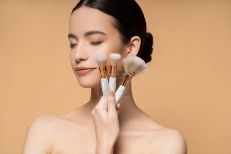 Junge asiatische Frau mit geschlossenen Augen und nackten Schultern hält Make-up-Pinsel isoliert auf beige