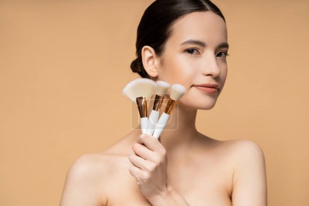 Téléchargez les photos : Brunette asiatique femme aux épaules nues tenant des pinceaux de maquillage isolés sur beige - en image libre de droit