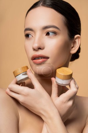 Pretty young asian woman with naked shoulders holding jars with cosmetic cream isolated on beige