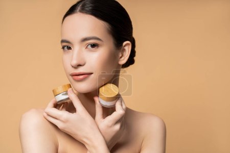 Joven mujer asiática con hombros desnudos sosteniendo frascos con crema cosmética y posando aislado en beige