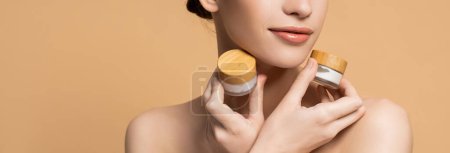 Cropped view of woman with naked shoulders holding jars of cosmetic cream isolated on beige, banner