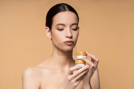Brunette asian woman with naked shoulders holding jars with cosmetic cream isolated on beige