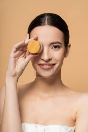 Cheerful asian woman in top with naked shoulders holding cosmetic cream near face isolated on beige