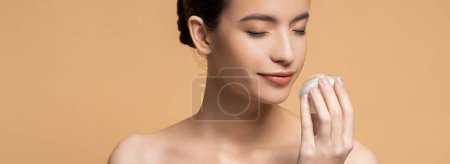 Young asian woman with naked shoulders smelling cosmetic cream isolated on beige, banner
