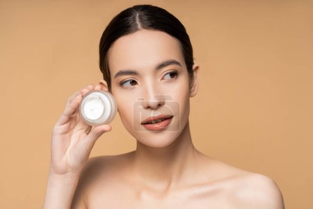 Young brunette woman with perfect skin and naked shoulders holding cosmetic cream isolated on beige
