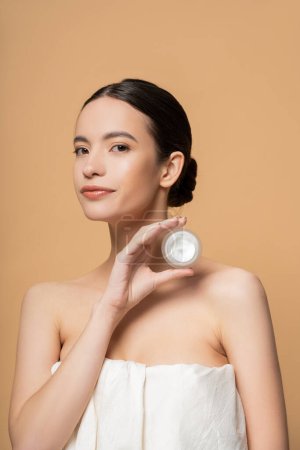 Young asian woman in top with naked shoulders holding cosmetic cream isolated on beige