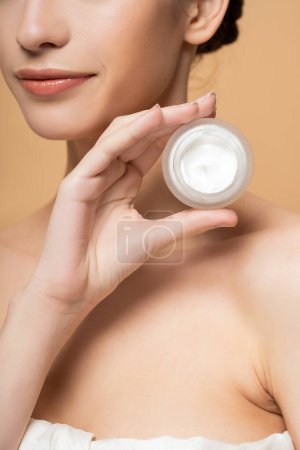 Cropped view of young woman in top holding cosmetic cream and standing isolated on beige