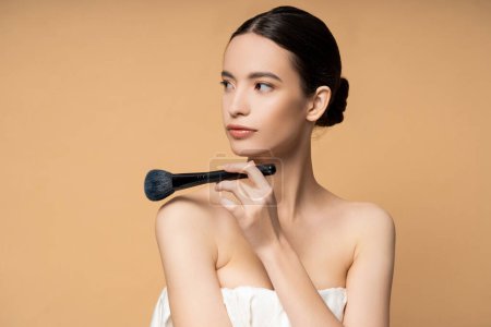 Young asian woman in top holding blush brush near naked shoulders isolated on beige