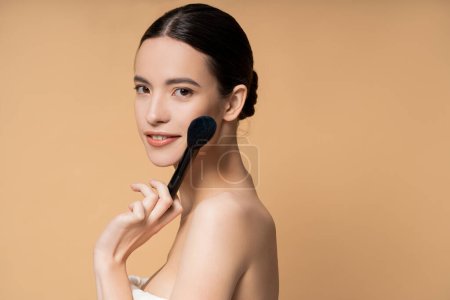 Smiling young asian woman with naked shoulders holding blush brush near face isolated on beige