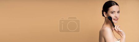 Photo for Young asian model with naked shoulder smiling and holding makeup brush isolated on beige, banner - Royalty Free Image