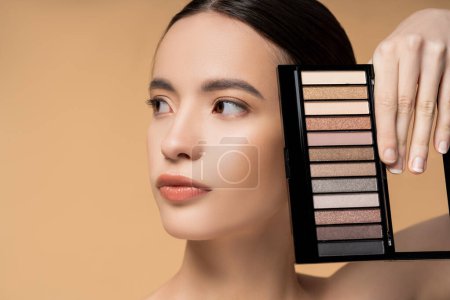 Portrait of young asian woman with natural makeup holding eyeshadow palette isolated on beige