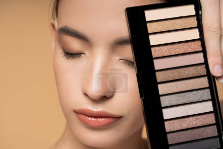 Close up view of asian woman with natural visage holding eyeshadow palette isolated on beige