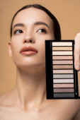 Young asian woman with natural makeup holding eyeshadow palette and looking away isolated on beige mug #666743588