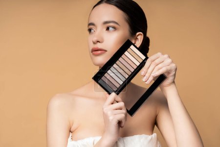 Brunette asian woman with perfect skin in top holding eyeshadow palette isolated on beige