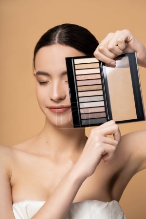 Young asian model with natural makeup in top covering face with eyeshadow palette isolated on beige