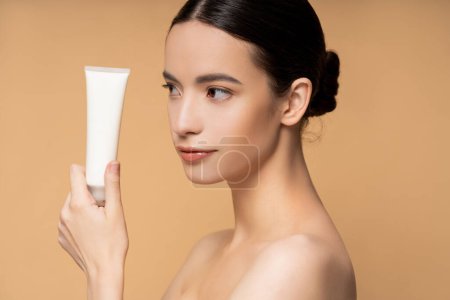 Young asian model with naked shoulders holding cosmetic lotion while posing isolated on beige