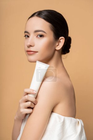 Photo for Pretty young asian model in top holding cosmetic lotion and looking at camera isolated on beige - Royalty Free Image