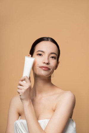Retrato de joven modelo asiático en la parte superior celebración de bálsamo cosmético y mirando a la cámara aislada en beige