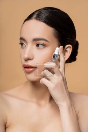 Pretty asian woman with naked shoulders holding cosmetic serum while posing isolated on beige