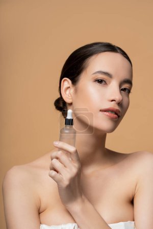 Asian woman with naked shoulders holding cosmetic serum and looking at camera isolated on beige