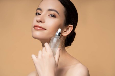 Pleased young asian woman with naked shoulders holding cosmetic serum isolated on beige