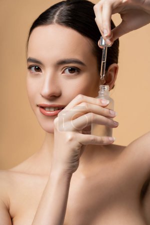 Portrait of confident and smiling asian woman with naked shoulders holding serum isolated on beige
