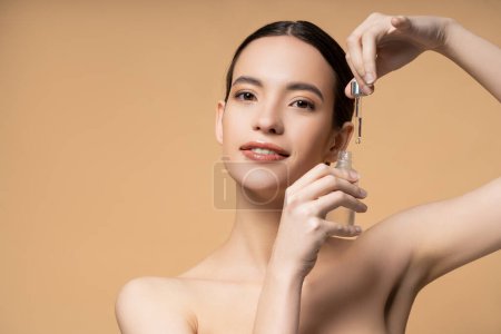 Positive asian woman with naked shoulders posing with cosmetic serum isolated on beige