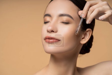 Young asian woman with closed eyes applying cosmetic serum while posing isolated on beige