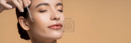 Mujer asiática bastante joven con los ojos cerrados aplicando suero cosmético aislado en beige, pancarta
