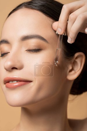 Vista de cerca de alegre joven modelo asiático aplicando suero cosmético aislado en beige