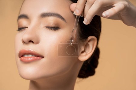Close up view of pleased asian woman applying cosmetic serum  while standing isolated on beige