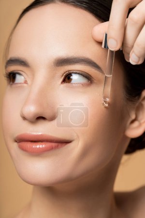 Close up view of young brunette asian woman applying cosmetic serum on cheek isolated on beige