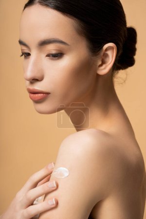 Portrait of young asian brunette woman applying cosmetic cream on shoulder isolated on beige