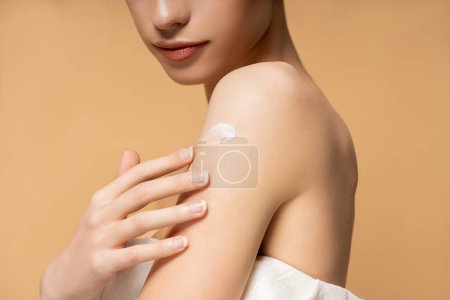 Cropped view of young woman in top with naked shoulder applying body cream isolated on beige