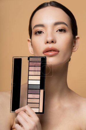 Confident young asian woman with naked shoulders holding eyeshadow palette isolated on beige