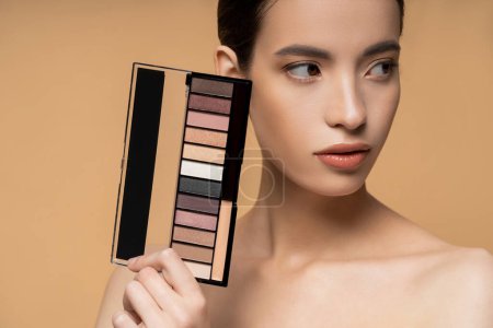 Asian woman with naked shoulders holding eyeshadow palette and looking away isolated on beige