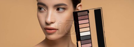 Pretty young asian woman with natural makeup holding eyeshadow palette isolated on beige, banner