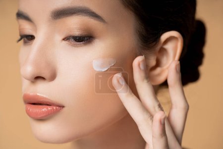 Nahaufnahme einer jungen asiatischen Frau, die Gesichtscreme auf die Wange aufträgt, isoliert auf beige