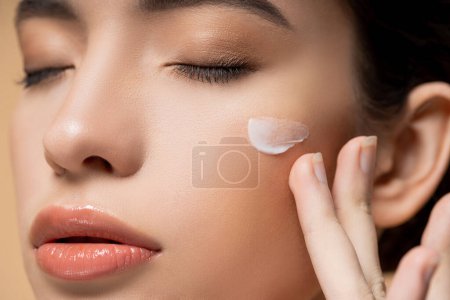 Vista de cerca de la joven mujer asiática con la piel perfecta aplicación de crema facial en la mejilla aislado en beige