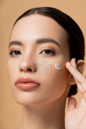 Retrato de mujer asiática joven aplicando crema facial en la mejilla y mirando a la cámara aislada en beige