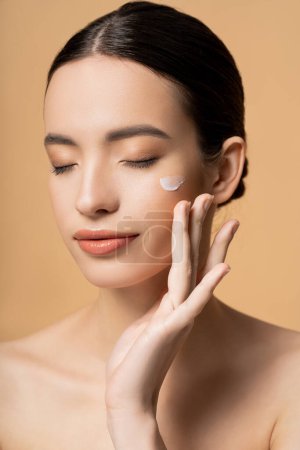 Young asian model with closed eyes applying face cream on cheek isolated on beige