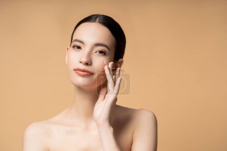Young asian woman looking at camera while applying face cream and standing isolated on beige