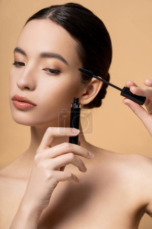 Young asian woman with naked shoulders applying mascara while standing isolated on beige