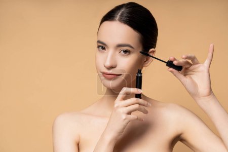 Junge asiatische Frau mit nackten Schultern hält Mascara und Applikator isoliert auf beige